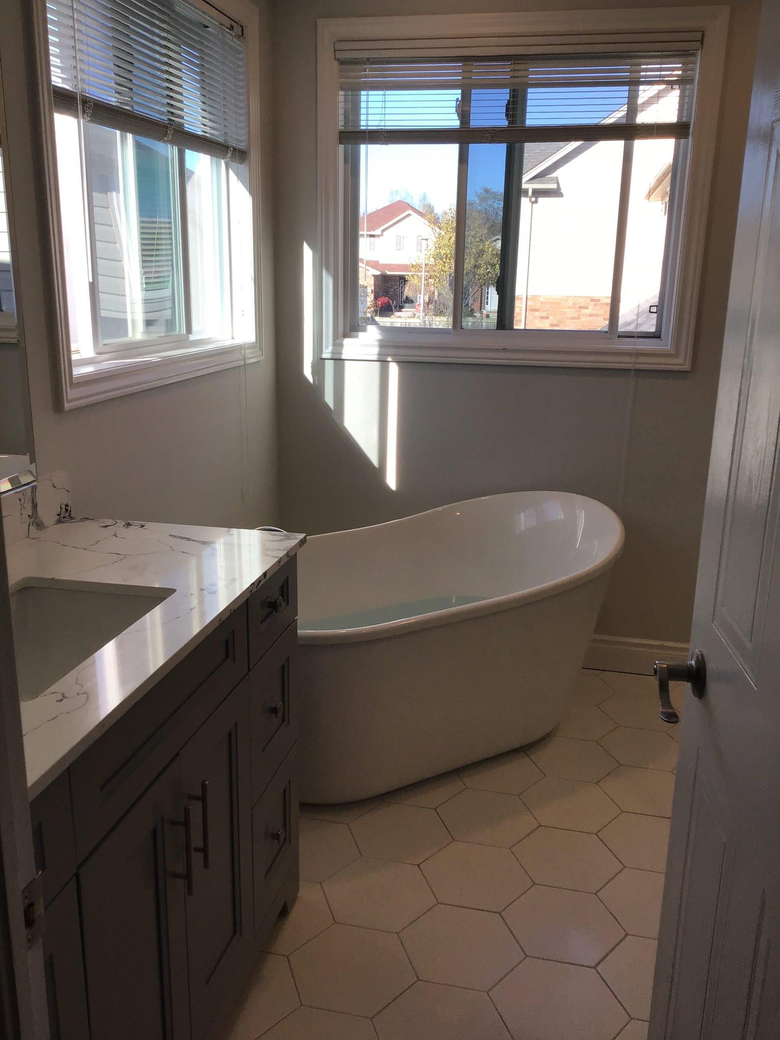 bathroom-renovation-london-ontario-donaldson.jpg A bathroom with a white freestanding tub, a gray vanity with marble top, hexagonal floor tiles, and two windows.