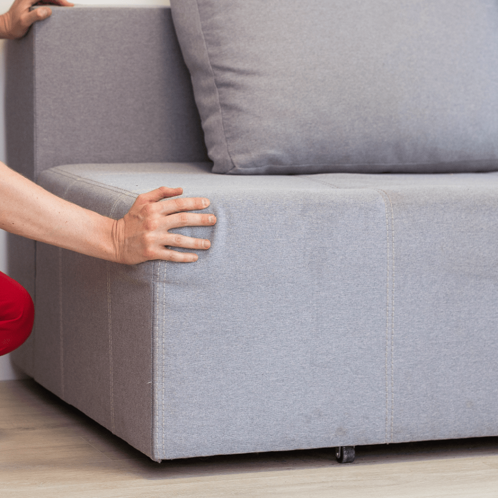 Person's hands on a grey sofa with a cushion and backrest. One hand on the seat edge, another on the backrest. Casters underneath.
